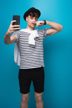 mavi arka planda yüzüne yakın çizilmiş bıyık lı parmağını tutarken akıllı telefonla selfie çeken yakışıklı adam
