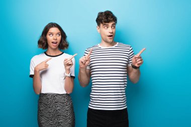 cheerful man and woman looking away and pointing with fingers on blue background clipart