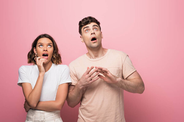 surprised man and woman gesturing while looking up on pink background