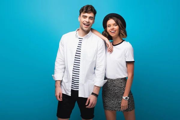 cheerful, trendy couple smiling at camera on blue background