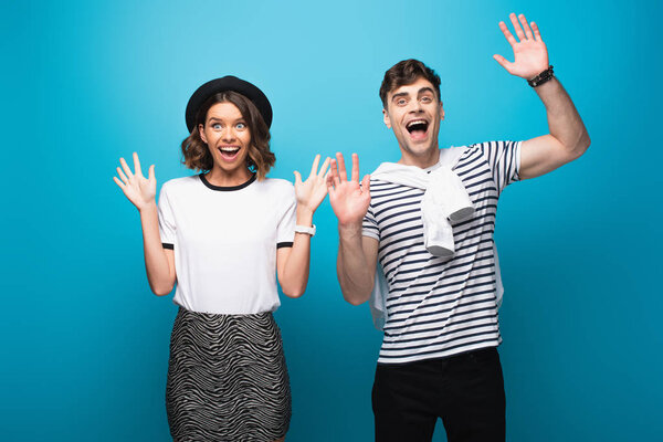 excited man and woman waving hands and smiling at camera on blue background