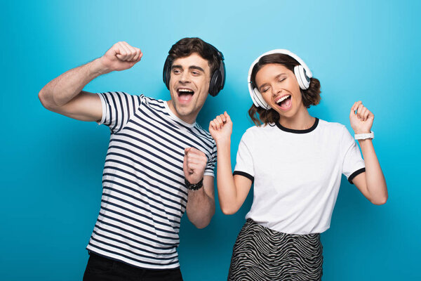 excited man and woman dancing while listening music in headphones on blue background