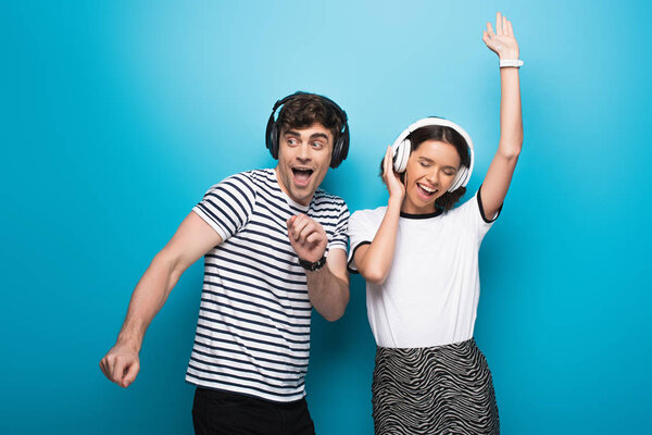 happy man and woman dancing while listening music in headphones on blue background