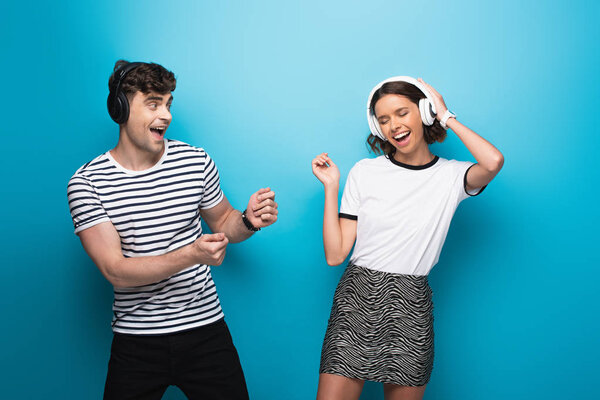 cheerful man and woman dancing while listening music in headphones on blue background
