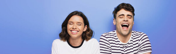 panoramic shot of cheerful man and woman laughing with closed eyes on blue background