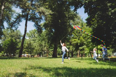 happy family running and playing with flying kite in park with copy space clipart