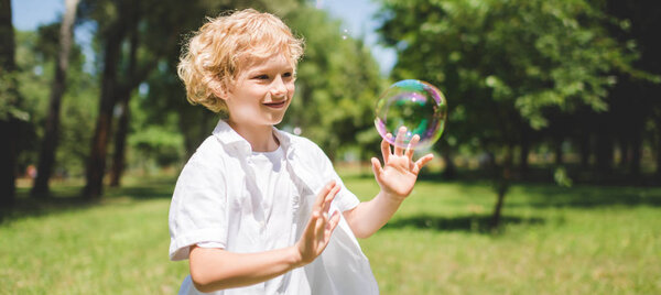 panoramic shot of cute boy Gesturing near soap bubbles in park