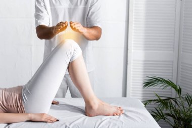 partial view of healer standing near woman on massage table and holding hands above her knees clipart
