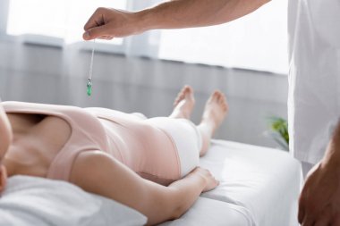 cropped view of hypnotist standing near woman on massage table and holding green stone clipart