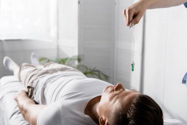 cropped view of hypnotist standing near man on massage table and holding green stone clipart