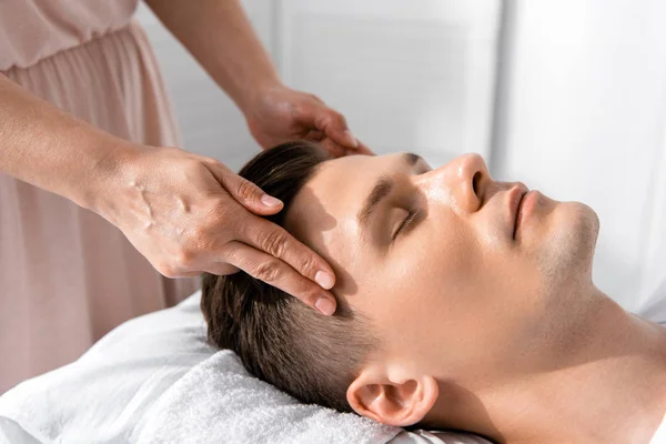 cropped view of masseur standing near man with closed eyes and touching his head