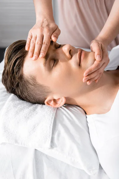 cropped view of masseur standing near man with closed eyes and touching his face