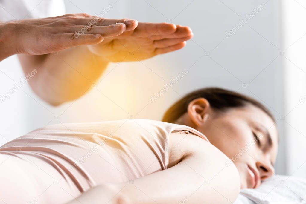 cropped view of healer standing near woman lying with closed eyes and holding hands above her body