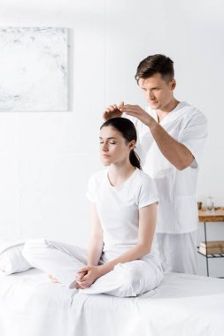 woman sitting on massage table with closed eyes while healer holding hands above her head clipart