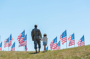Amerikan bayrakları yakınında kızı ile el ele tutuşan askeri üniformalı adamın geri görünümü 