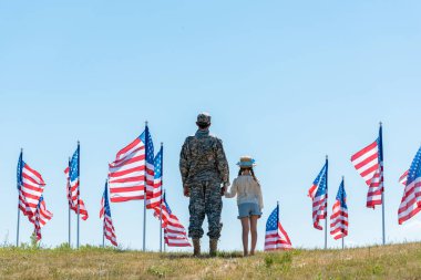 back view of military man in uniform holding hands with daughter near american flags  clipart