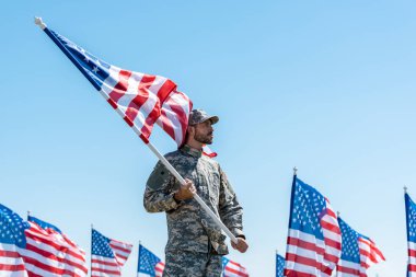 mavi gökyüzüne karşı dururken askeri üniforma ve şapka amerikan bayrağı tutan yakışıklı adam 