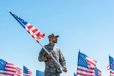 mavi gökyüzüne karşı dururken askeri üniforma ve şapka amerikan bayrağı tutan yakışıklı asker 