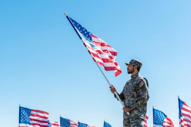 mavi gökyüzüne karşı dururken Amerikan bayrağı tutan üniformalı yakışıklı askeri adam 