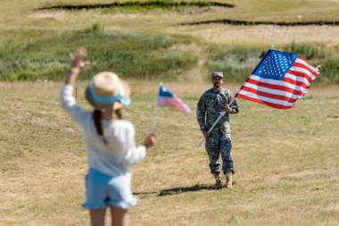 askeri üniformalı adam yakınında Amerikan bayrağı tutarken el sallayarak hasır şapkalı çocuğun arka görünümü 