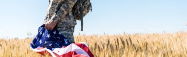 panoramic shot of patriotic soldier in military uniform holding american flag while standing in field  clipart