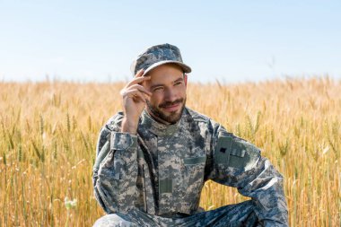 positive military man touching cap and smiling in field  clipart