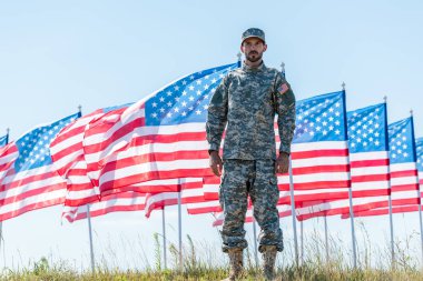 amerikan bayrakları ve mavi gökyüzü yakınında duran üniformalı yakışıklı asker 