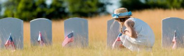 mezarlıkta Amerikan bayrağı ile mezar taşı yakınında oturan elbise ve hasır şapka lı çocuğun panoramik görüntüsü 