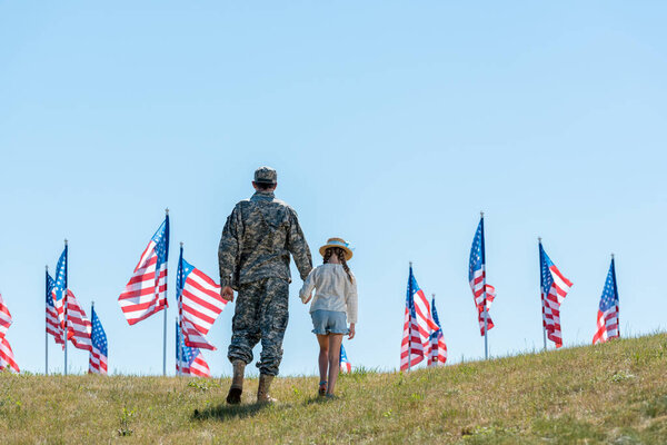 back view of father in military uniform holding hands with daughter near american flags 