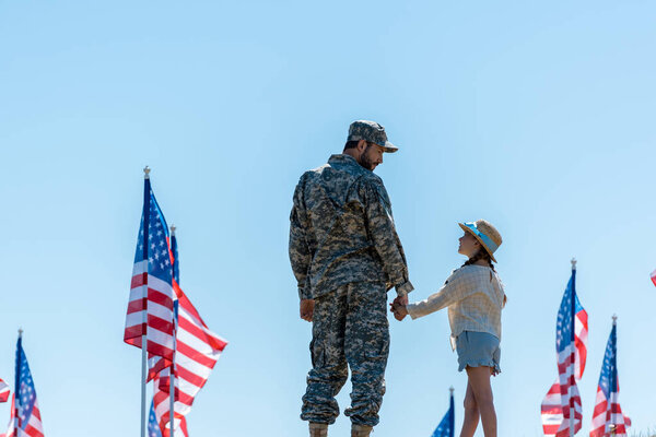 father in military uniform holding hands with cute daughter near american flags 