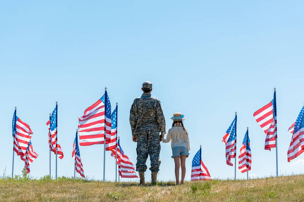 back view of military man in uniform holding hands with daughter near american flags 