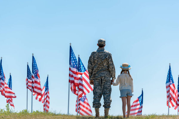 back view of father in military uniform holding hands with child near american flags 