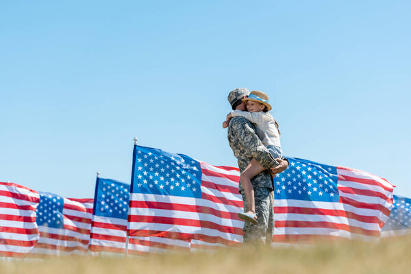 selective focus of military man holding in arms cheerful kid near american flags 