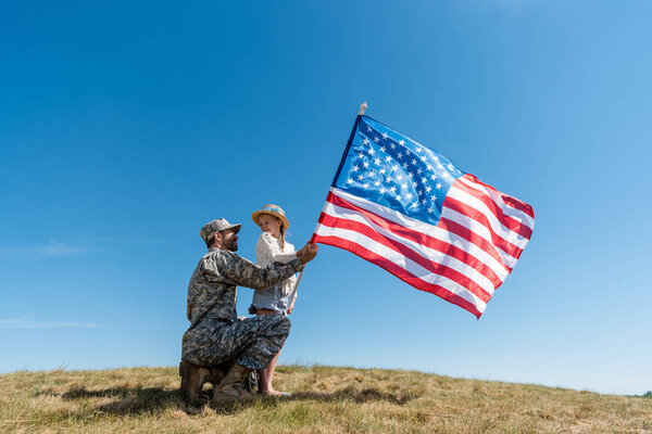 father in military uniform and happy kid holding american flag against sky 