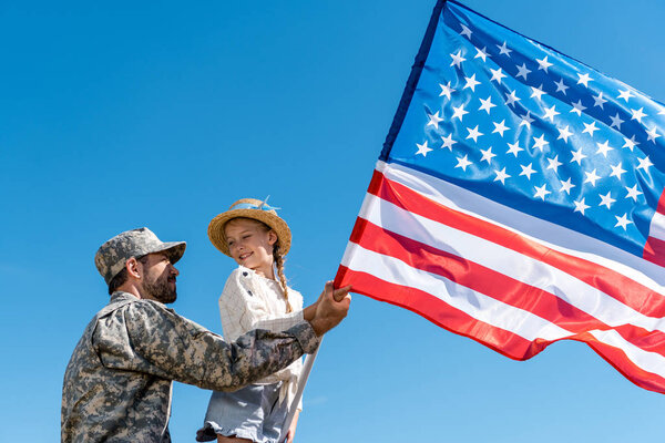 low angle view of father in military uniform and happy kid holding american flag against sky 