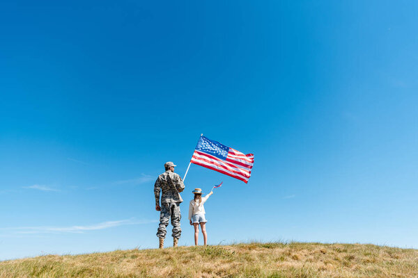 back view of kid in straw hat and military father holding american flags 