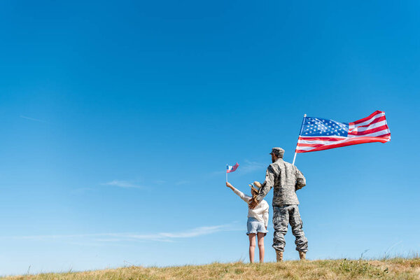 back view of child in straw hat and military father holding american flags while standing on grass 