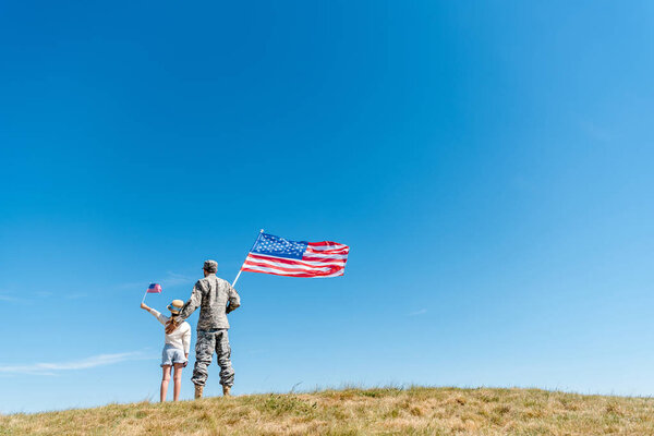 Вид сзади ребенка в соломенной шляпе и военного, держащего в руках американские флаги
