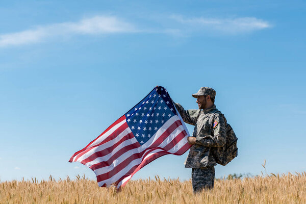 happy soldier in military uniform standing in field with golden wheat and holding american flag 