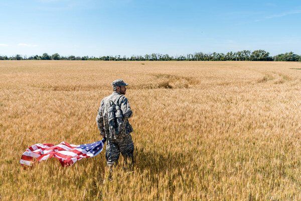 handsome man in military uniform holding american flag while standing in field with wheat 