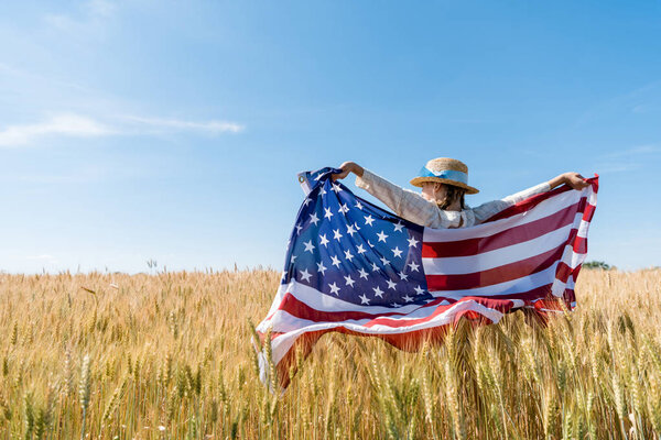 back view of kid in straw hat holding american flag in golden field 