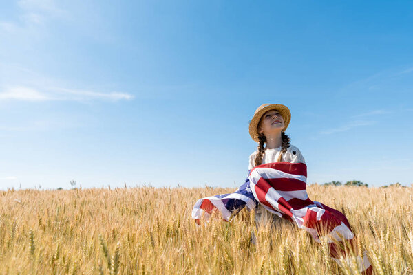 smiling kid in straw hat holding american flag in golden field with wheat 