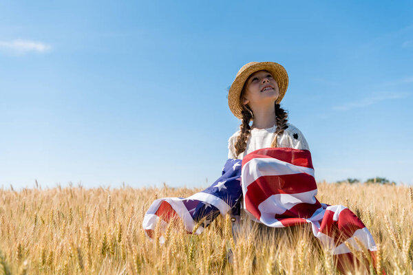 smiling child in straw hat holding american flag in golden field with wheat 
