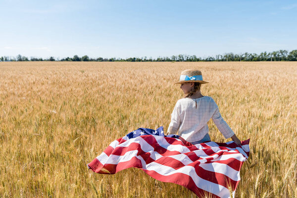 cute child in straw hat holding american flag in golden field with wheat 
