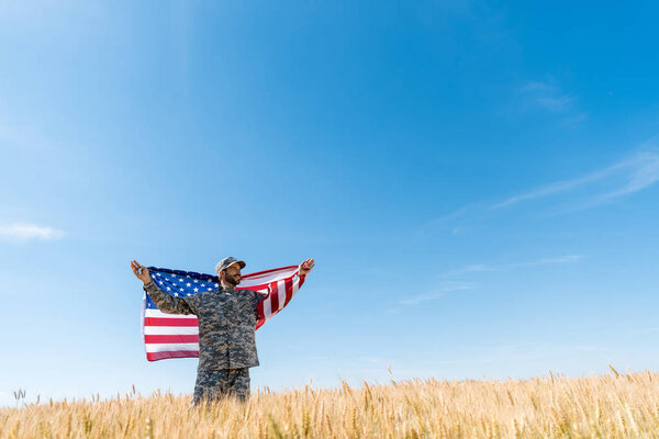 soldier in cap and uniform holding american flag in field with wheat 