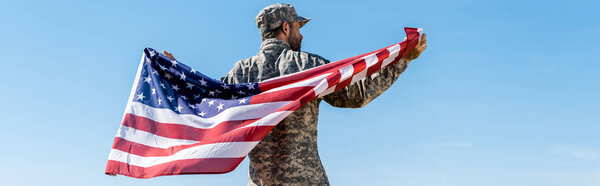 panoramic shot of soldier in cap and uniform holding american flag against blue sky 