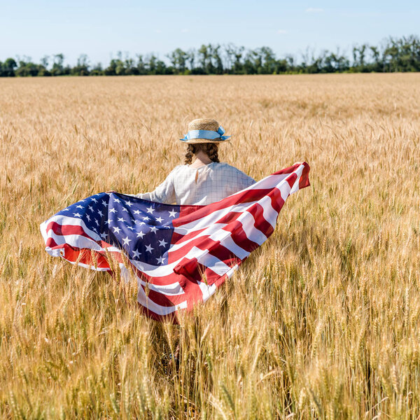 back view of child in straw hat holding american flag in golden field 