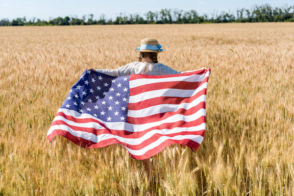 back view of kid in straw hat holding american flag in golden field 