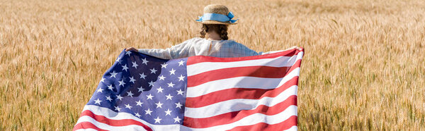 panoramic shot of kid in straw hat holding american flag with stars and stripes in golden field 