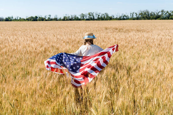 back view of kid holding american flag with stars and stripes in golden field 